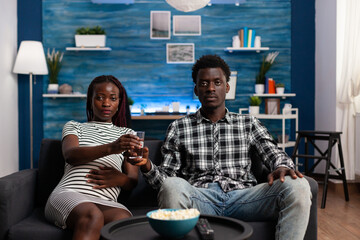 Young african american couple expecting baby and watching TV while man offers pregnant woman a glass of water. People sitting on sofa and watching television programs together at home.