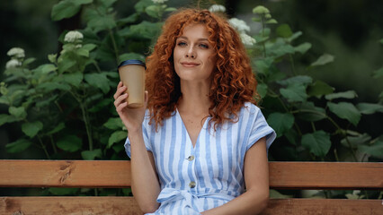 happy redhead woman in dress holding paper cup while sitting on bench in park.