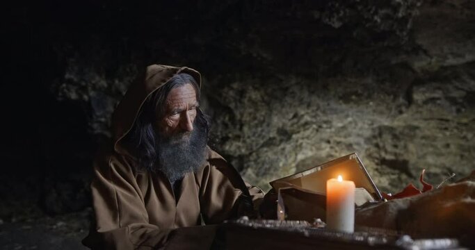 Senior monk in hood sitting in dark rocky cave with burning candle and taking notes with quill