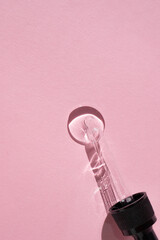 Glass pipette and a round drop of essence on a pink background. Vertical image, flat lay, space for text.