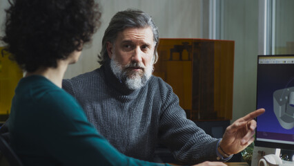 Mature and young men in casual clothes pointing at computer monitor and discussing 3D model during work in contemporary studio