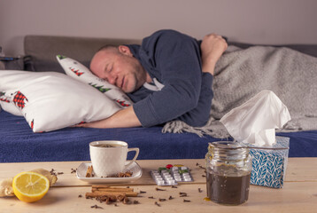 Mężczyzna przeziębiony leży z gorączką w łóżku na pierwszym planie widzimy tabletki, herbatę, miód, cytrynę i korzenie pomocne przy leczeniu. - obrazy, fototapety, plakaty