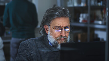 Middle aged bearded man in glasses creating 3D model on computer while sitting behind glass in...