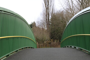 Green metal bridge with yellow handrail leading to a forested area in Manchester UK - 484164477
