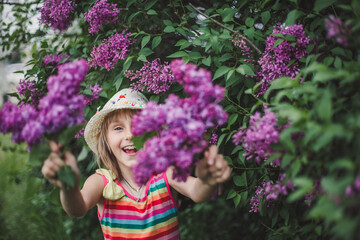 Laughing European brunette girl in a beige hat and multicolored striped dress holds lilac flowers in her hands