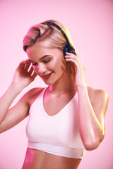 Obraz na płótnie Canvas Enjoying music. Close up of cute and sporty woman in headphones listening music and smiling while standing against pink background