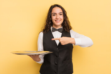 young pretty hispanic woman smiling cheerfully, feeling happy and pointing to the side. waiter and...