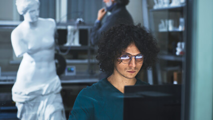 Young man in glasses with curly hair creating 3D model on computer for printing while sitting...