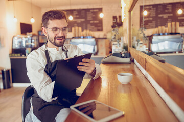 Fototapeta premium Joyful male barista writing on clipboard in cafe