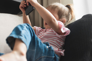 A little girl is sitting on the couch and holding a smartphone in her outstretched hands. Selective focus. Close-up.