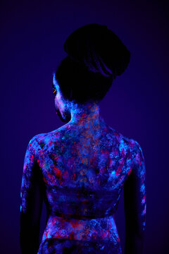 slim black female with luminescence, fluorescence, body art isolated on dark background. rear view on young woman looking charming, in ultraviolet light. extraterrestrial fantastic shoot