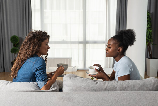 Two sad depressed and anxious diverse women talking at home. Female friends supporting each other while they drinking coffee or tea. Problems, friendship and relationships difficulties care concept