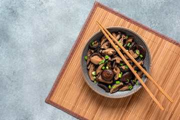 Fried shiitake mushrooms with green onions in a bowl on a bamboo mat. Gray rustic background. Asian vegan food. Top view.