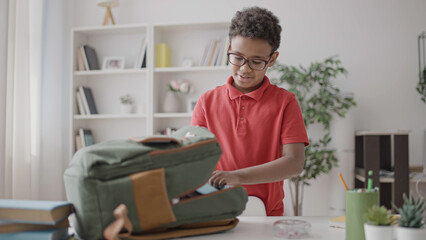 Pupil in eyeglasses packing backpack for school, putting books inside, routine