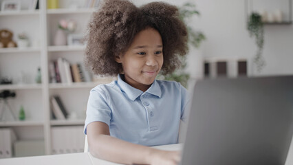 Happy school girl typing on laptop at home during online lesson, e-learning