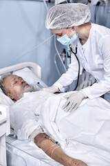 Senior Caucasian Male Patient receiving consultation from doctor using stethoscope for heartbeat exam. Confident Female Nurse consulting retired man with disease, in modern hospital ward