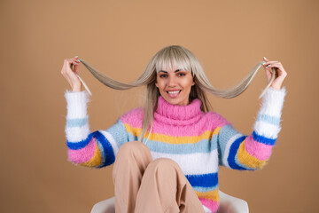 A blonde with long hair and bangs, with nude makeup and in a multi-colored bright fluffy sweater on a beige background smiles cheerfully