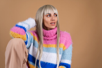 A blonde with long hair and bangs, with nude makeup and in a multi-colored bright fluffy sweater on a beige background smiles cheerfully