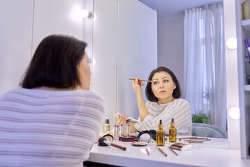 Fototapeta premium Beautiful middle aged woman doing makeup in front of a mirror.