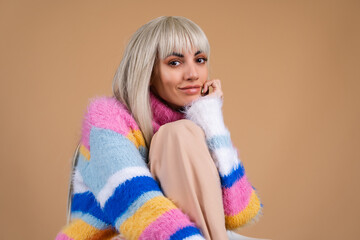 A blonde with long hair and bangs, with nude makeup and in a multi-colored bright fluffy sweater on a beige background smiles cheerfully