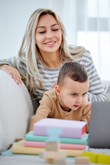 caucasian blonde mom and little boy sitting on sofa at home watch video on modern laptop,caucasian beautiful mother or nanny relax with small kid boy child using computer, in bright cozy room