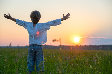 Happy boy with raised hands looks at the sunset  in summer. Boy in a field with his hands raised...