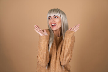 Blonde girl with bangs and brown daytime make-up in a sweater on a beige background shocked...
