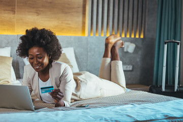 The woman lies on the bed with a laptop and a credit card and enters the data for the purchase. An...