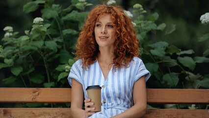 curly redhead woman in dress smiling and holding paper cup while sitting on bench in park.