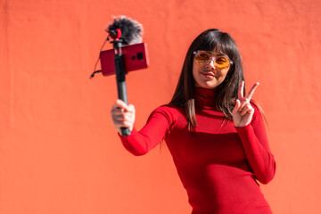 Brunette girl making the victory sign recording a video blog with the mobile phone and microphone,...