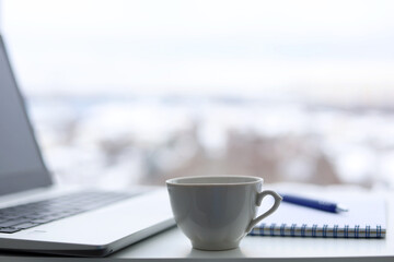 Tea cup, laptop and notepad with pen on a table against the window, view to winter city. Cozy workplace in home office