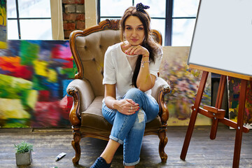 Portrait of attractive female painter resting in comfortable vintage armchair and posing during weekend pastime in art gallery, Caucasian artist in casual clothes looking at camera indoors