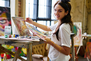 Portrait of cheerful female mastery with palette for mixing colors smiling at camera during time for dye creativity in art studio, happy Caucaisan woman posing while drawing abstract paintings