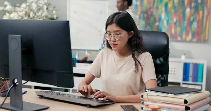 Nerd girl in big glasses and tshirt sits in comfortable chair in front of desk, taps fingers on computer keyboard, sends messages, emails, writes back, runs company website, office life, corporate