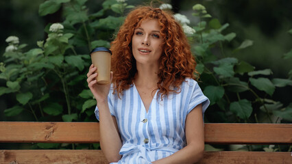 Naklejka premium pleased redhead woman in dress holding paper cup while sitting on bench in park.