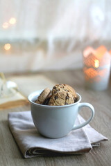 Fototapeta premium Cup of chocolate chip cookies, open book and reading glasses on the table. Lit candle and bokeh lights in the background. Hygge at home. Selective focus.