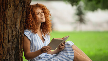 pleased redhead woman in striped dress holding book and sitting under tree trunk.