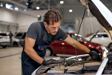 young focused american or caucasian male can fix anything. handsome car mechanic in overall uniform checking the engine under hood in modern clean car service station, workshop.