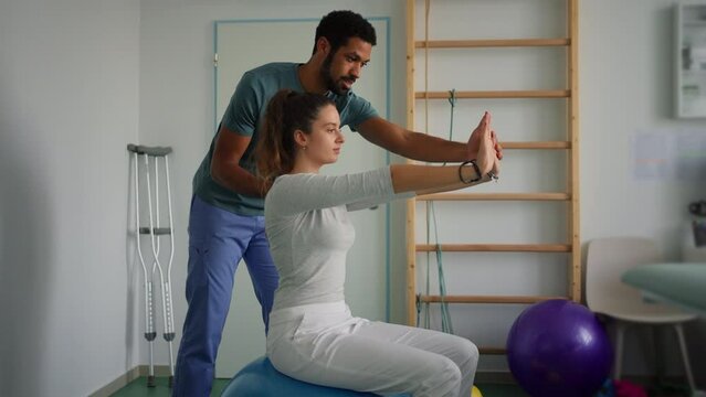 Young male physiotherapist exercising with young woman patient on ball in a physic room