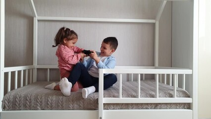 Two Children brother and sister are sitting in a Montessori bed and fighting for playing with gamepad Video games. 