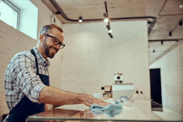Cheerful male bartender cleaning glass showcase in cafe