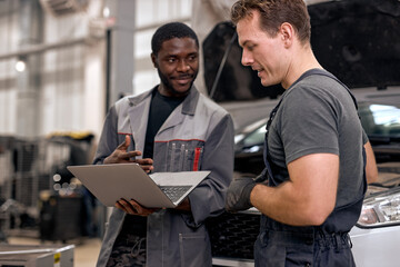 Naklejka na ściany i meble Handsome diverse mechanics in uniform examining car, using laptop while working in auto service. black and caucasian Auto mechanics checking car,service via online insurance system at garage
