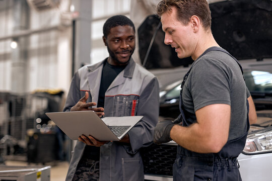 Handsome diverse mechanics in uniform examining car, using laptop while working in auto service. black and caucasian Auto mechanics checking car,service via online insurance system at garage