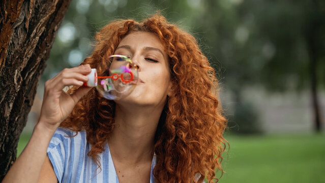 curly redhead woman blowing soap bubble in park.