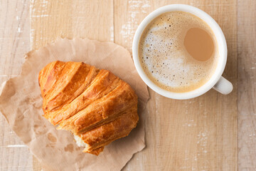 White ceramic cup of coffee latte and half of croissant on parchment paper, wooden table background, top view