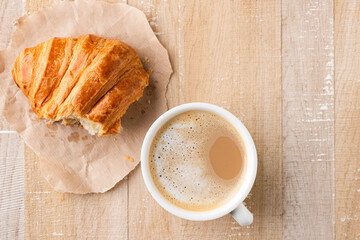 White ceramic cup of coffee latte and half of croissant on parchment paper, wooden table background, top view