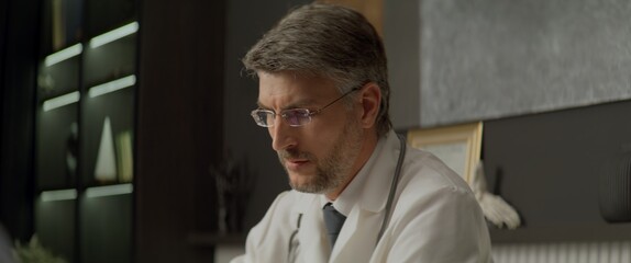 Fototapeta premium Portrait of Caucasian doctor delivering telemedicine consultation to a patient from his office. Telehealth, telemedicine, remote consultation service. Coronavirus, COVID-19