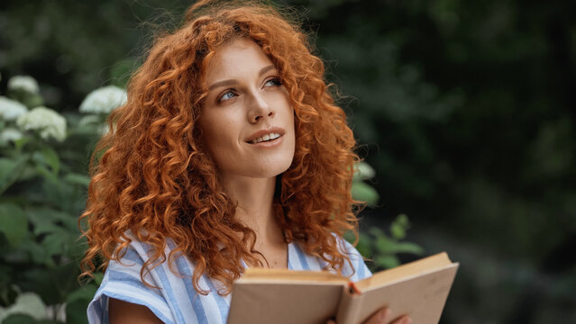 dreamy redhead woman smiling while holding book outdoors.