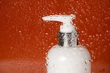 Naklejka na ściany i meble White unbranded dispenser bottle behind glass with water drops on orange background, cosmetics