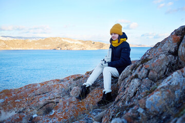 A happy girl traveler is sitting on a rock near Lake Baikal, holding a cup of hot tea from a thermos in her hand. Winter landscape. Freedom, the concept of travel, active recreation. 